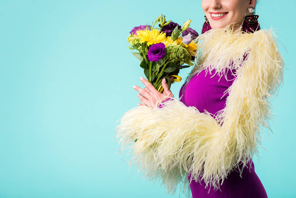 μερική άποψη του χαμογελαστό κορίτσι κόμμα σε μωβ φόρεμα με φτερά κρατώντας μπουκέτο λουλούδια απομονωμένα σε τυρκουάζ  - Φωτογραφία, εικόνα