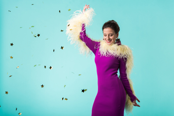 happy party girl in purple dress with feathers dancing under falling confetti isolated on turquoise  - Photo, image
