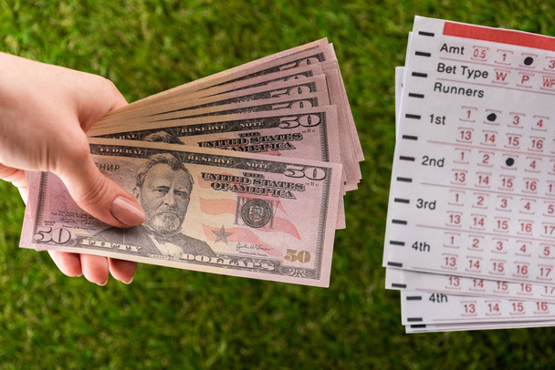 cropped view of woman holding dollar banknotes near betting lists on green grass, sports betting concept - Φωτογραφία, εικόνα