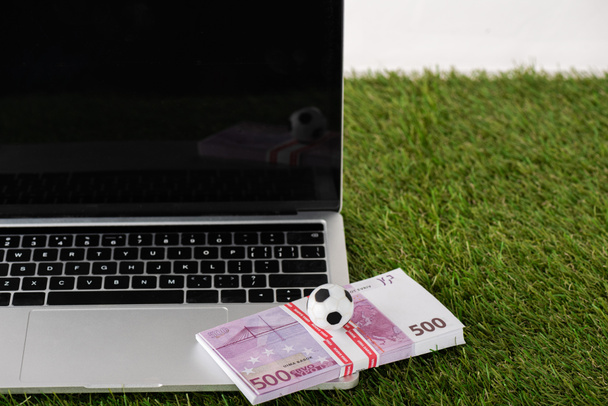 speelgoed voetbal en eurobankbiljetten op laptop met blanco scherm op groen gras geïsoleerd op wit, sportweddenschappen concept - Foto, afbeelding