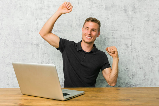若い起業家は、特別な日を祝う机の上で彼のラップトップで作業し、ジャンプし、エネルギーで腕を上げます. - 写真・画像