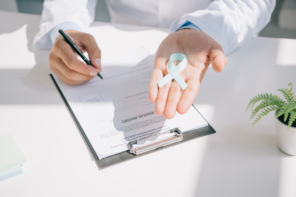 vista cortada de médico segurando fita de consciência azul enquanto escrevendo no relatório médico
 - Foto, Imagem