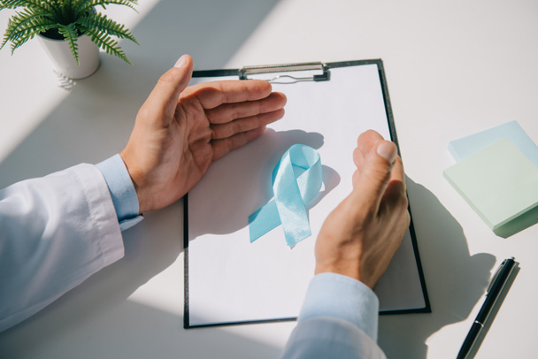 částečný pohled na lékaře držícího se za ruce poblíž modré informační stuhy na schránce s prázdným papírem - Fotografie, Obrázek