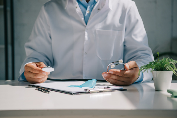 przycięty widok lekarza siedzącego przy biurku i trzymającego stetoskop blisko niebieskiej wstążki świadomości - Zdjęcie, obraz