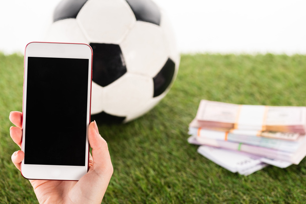 vista parcial de la mano femenina con smartphone cerca de paquetes de dinero y pelota de fútbol sobre hierba verde aislado en blanco, concepto de apuestas deportivas
 - Foto, Imagen