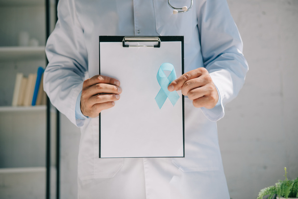 vue recadrée du médecin tenant le presse-papiers avec du papier vierge et un ruban de sensibilisation bleu
 - Photo, image