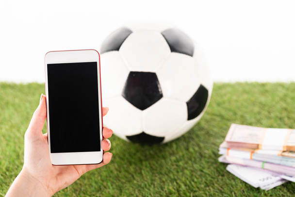 vista recortada de la mano femenina con teléfono inteligente cerca de paquetes de dinero y pelota de fútbol sobre hierba verde aislado en blanco, concepto de apuestas deportivas
 - Foto, Imagen