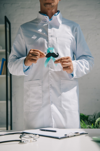 částečný pohled na lékaře v bílém plášti držícího modrou informační stuhu s papírovým knírem - Fotografie, Obrázek
