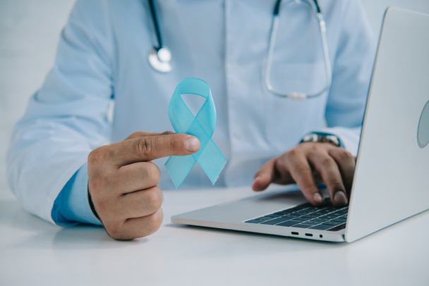 cropped view of doctor holding blue awareness ribbon while using laptop - Фото, изображение
