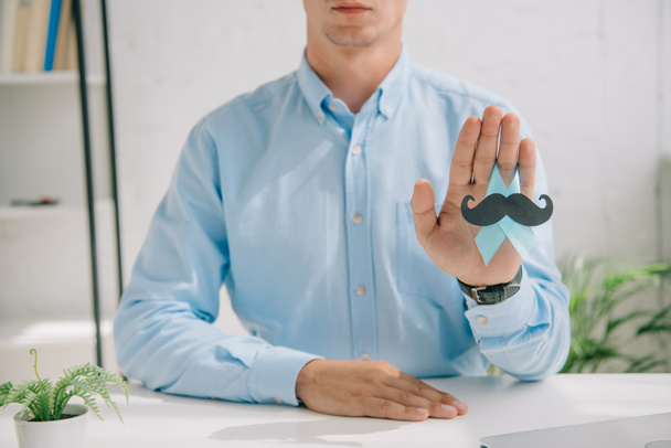 partial view of man in blue shirt holding blue awareness ribbon with paper cut mustache  - Фото, изображение