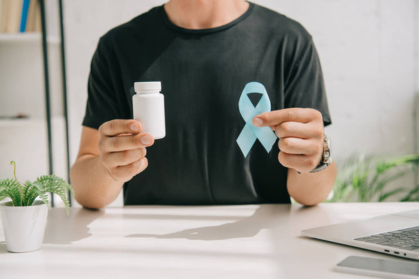 cropped view of man in black t-shirt holding blue awareness ribbon and container with pills - Photo, Image