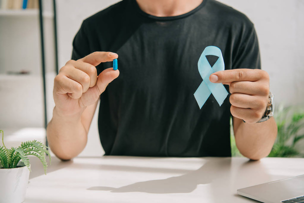 cropped view of man in black t-shirt holding blue awareness ribbon and pill - Φωτογραφία, εικόνα