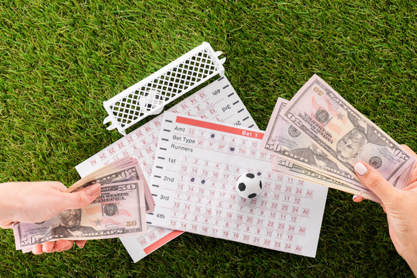částečný pohled na ženské ruce s dolarovými bankovkami v blízkosti fotbalových míčků, branek a sázkových listů na zelenou trávu, koncept sportovního sázení - Fotografie, Obrázek