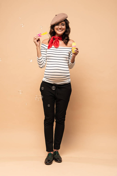 pregnant french woman in beret holding bottle with soap bubbles on beige background  - Foto, imagen