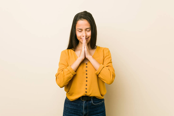 Młoda kobieta Hiszpanie na beżowym tle trzymając ręce w modlitwie w pobliżu ust, czuje się pewnie. - Zdjęcie, obraz