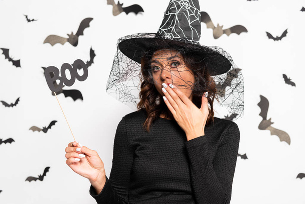 wstrząśnięta kobieta w kapeluszu czarownicy trzymając kij z literami boo w Halloween - Zdjęcie, obraz