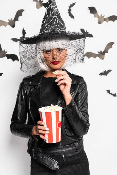 atractiva mujer en sombrero de bruja y peluca sosteniendo palomitas de maíz en Halloween
 - Foto, Imagen