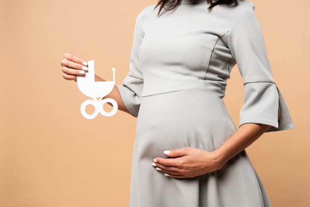 Ausgeschnittene Ansicht einer schwangeren Frau im grauen Kleid mit einem Kinderwagen aus Papier auf beigem Hintergrund  - Foto, Bild