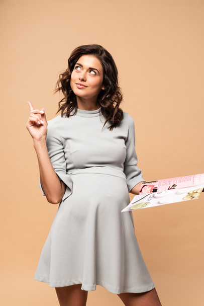 pregnant woman in grey dress holding magazine and showing idea gesture on beige background  - Photo, Image