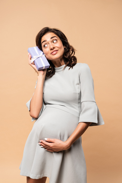 pregnant woman in grey dress holding gift and touching belly on beige background - Fotografie, Obrázek
