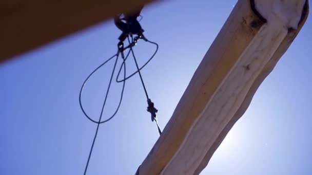 Crane with log for construction of wooden house. Clip. Suspended timber on crane ropes weighs on blue sky background with sun rays - Footage, Video