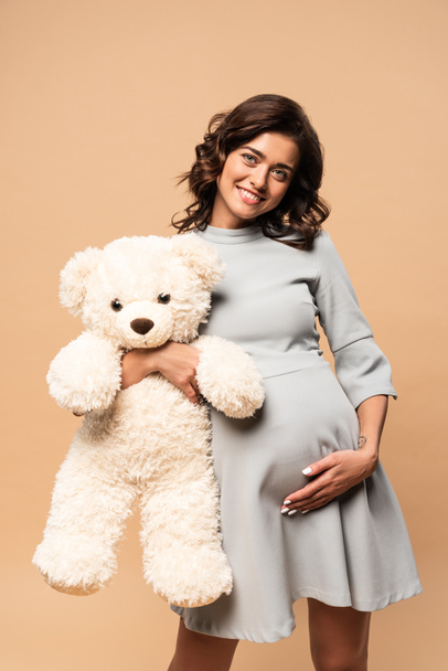 pregnant woman in grey dress holding teddy bear on beige background  - Фото, изображение