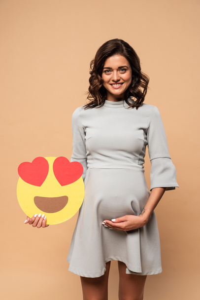 KYIV, UKRAINE - AUGUST 1, 2019: pregnant woman in grey dress holding smiley with heart eyes on beige background  - Foto, Imagem