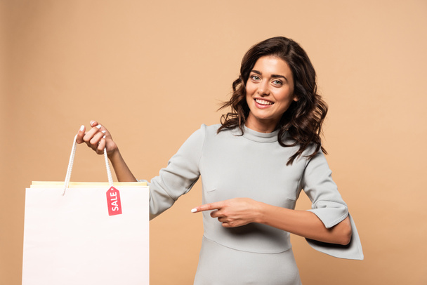 mujer embarazada en vestido gris apuntando con el dedo a la bolsa de la compra sobre fondo beige
  - Foto, Imagen
