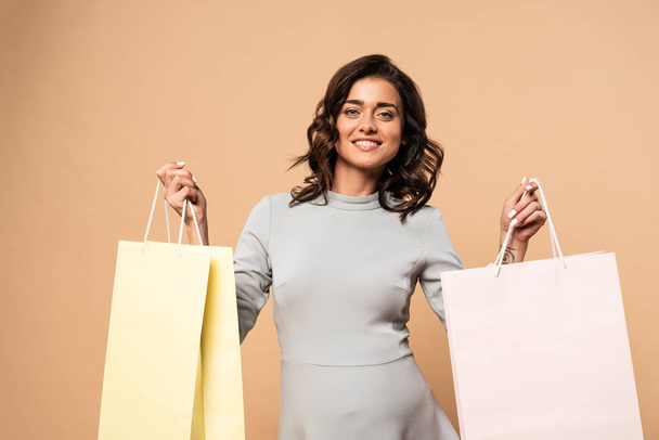 pregnant woman in grey dress smiling and holding shopping bags on beige background  - Φωτογραφία, εικόνα