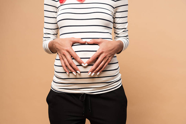 cropped view of pregnant woman showing heart sign on beige background  - Fotoğraf, Görsel