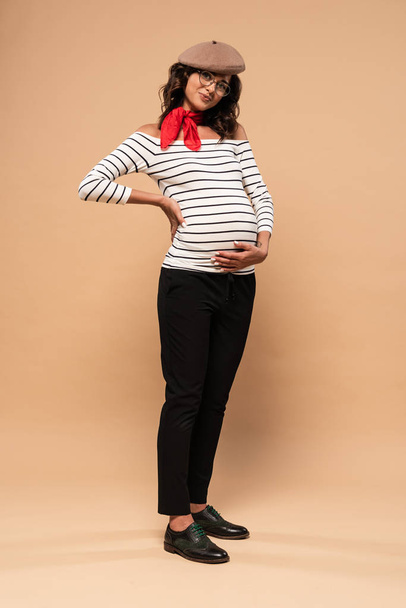 těhotná francouzky žena v baretu při pohledu na kameru na béžovém pozadí  - Fotografie, Obrázek