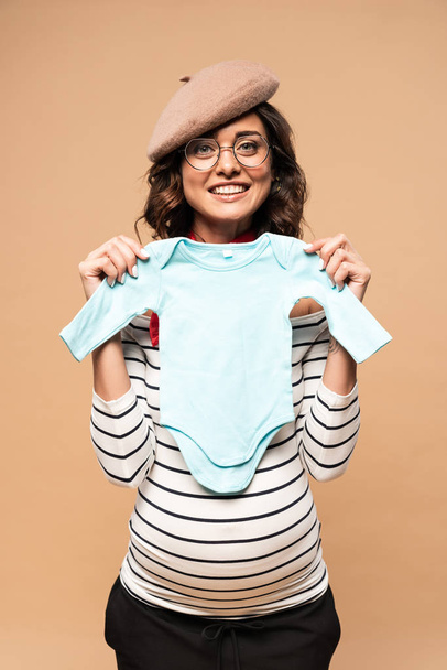 zwanger frans vrouw in baret glimlachen en het houden van overalls geïsoleerd op beige  - Foto, afbeelding