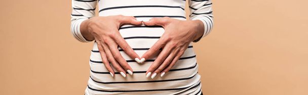 plano panorámico de la mujer embarazada mostrando el signo del corazón aislado en beige
 - Foto, imagen