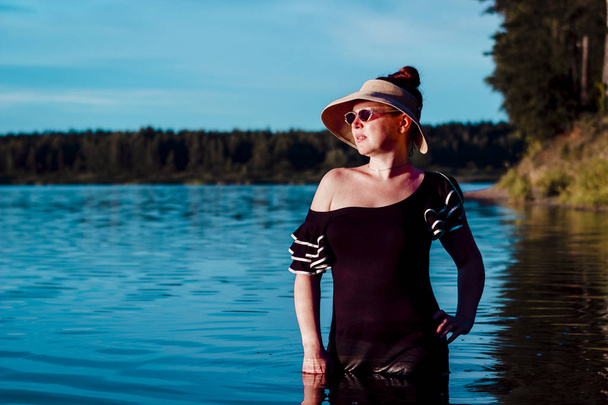麦わら帽子をかぶった少女は湖にいる。黒いドレスは水から濡れた。遠くに見える。肩裸. - 写真・画像