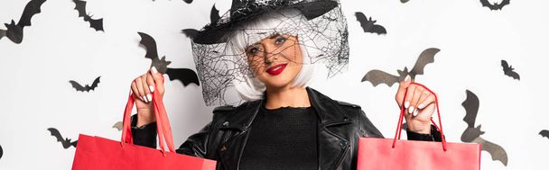 panoramic shot of attractive woman in witch hat and wig holding shopping bags in Halloween - Photo, Image