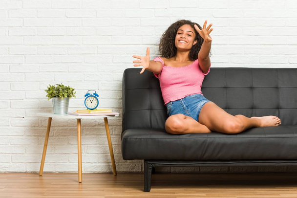 Молодая африканская американка, сидящая на диване, чувствует себя уверенно, обнимая камеру.
. - Фото, изображение