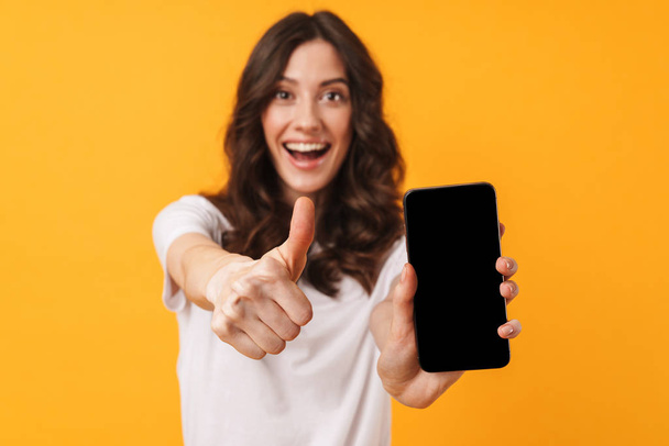Happy positive smiling young woman posing isolated over yellow wall background using mobile phone showing display and thumbs up. - Photo, image