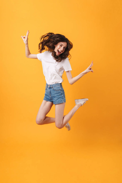 Emotional happy young woman jumping isolated over yellow wall background showing peace. - Foto, Imagem