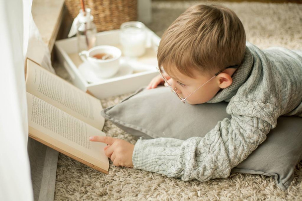 Красивый маленький мальчик в вязаном свитере читает у окна кружку горячего чая. Уютно. Осень
. - Фото, изображение