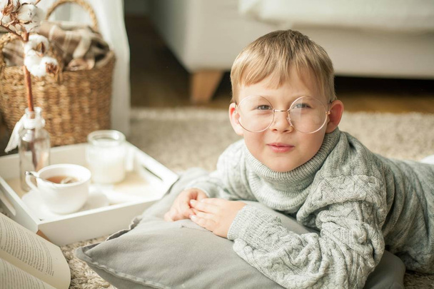 A beautiful little boy in a knitted sweater is reading by the window with a mug of hot tea. Cozy. Autumn. - Foto, imagen