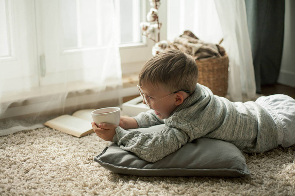A beautiful little boy in a knitted sweater is reading by the window with a mug of hot tea. Cozy. Autumn. - Foto, Imagen