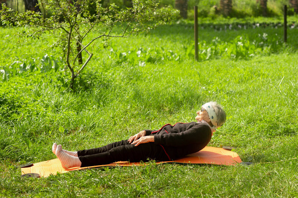 Μια ηλικιωμένη γυναίκα 90 ετών που κάνει γυμναστική στον κήπο πάνω σε στρώμα γιόγκα, το πρωί του καλοκαιριού. Έννοια της μακροβιότητας - Φωτογραφία, εικόνα