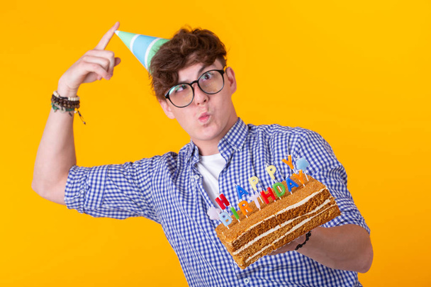 Portrait of a funny positive guy with a paper cap and glasses holding a congratulatory cake in his hands on a yellow background. Concept and fun and celebration. Advertising space. - Φωτογραφία, εικόνα