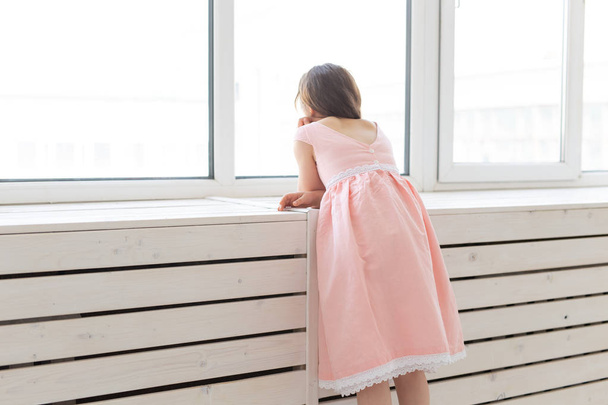Charming little girl in a lush peach princess dress looks out the window of her room. The concept of childrens desires and expectations of a miracle. - 写真・画像