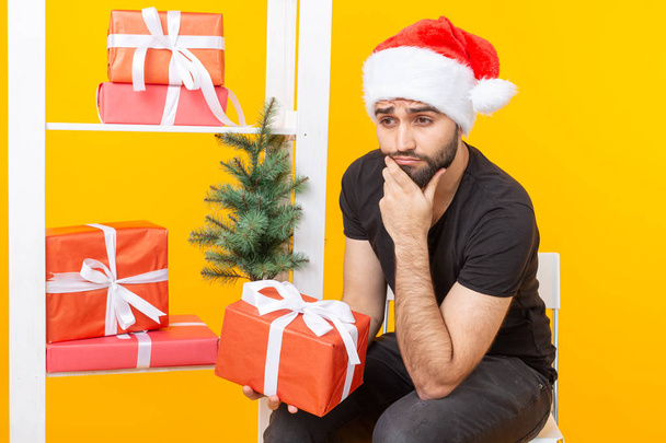 Young handsome man in a Santa Claus hat is holding congratulatory gifts next to a Christmas tree. Concept of holidays of christmas and new year. - Foto, Bild