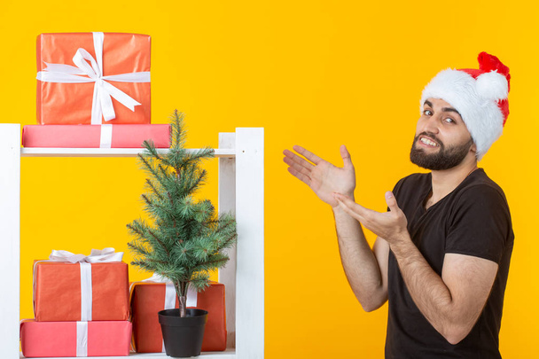 あごひげとサンタクロースの帽子をかぶった若いポジティブな男性は、黄色の背景に贈り物やクリスマスツリーとブースの近くに立っています。クリスマスと新年のお祝いの贈り物や割引の概念. - 写真・画像