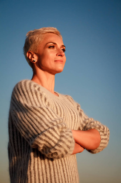 erwachsene blonde Frau mittleren Alters im Pullover steht vor blauem Himmel am Strand des Sonnenuntergangs, selektiver Fokus - Foto, Bild