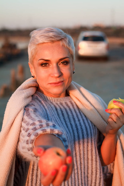 Adult middle-aged blonde woman in a sweater eating an apple and holding out another one to the camera, selective focus - Photo, Image