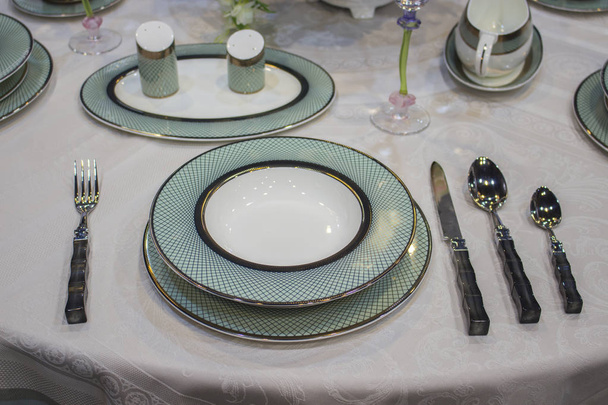Beautiful turquoise blue white plates with gold on the tablecloth. Festive table setting deep and flat plates knife fork spoons. Beautiful dishes - Photo, Image