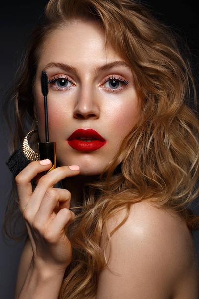 Menina bonita com lábios vermelhos e maquiagem clássica e cachos com rímel na mão. Cara de beleza
 - Foto, Imagem